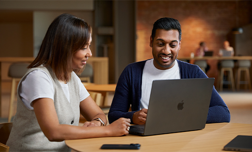 Amar ทำงานร่วมกับเพื่อนร่วมงานขณะนั่งอยู่กับ MacBook ของตัวเองที่โต๊ะในพื้นที่ส่วนกลาง
