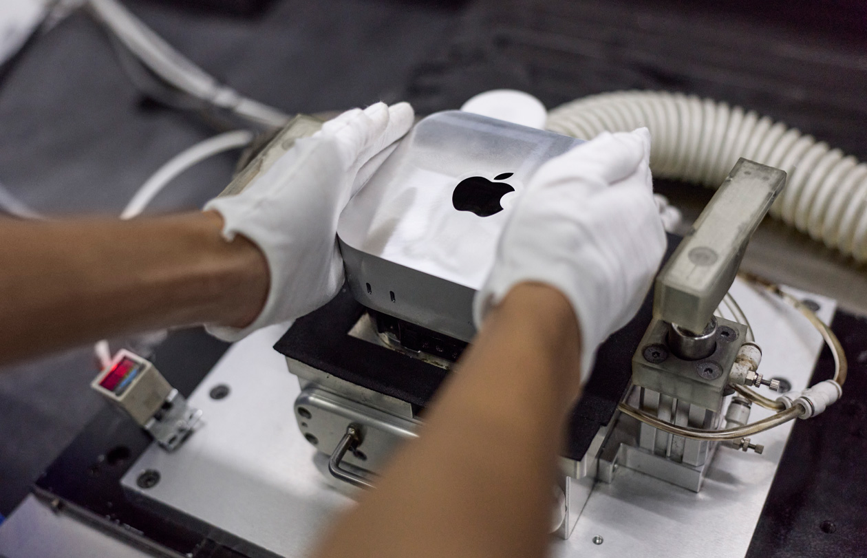 มือที่สวมถุงมือวาง Mac mini ลงในเครื่องจักรผลิตระหว่างการประกอบ