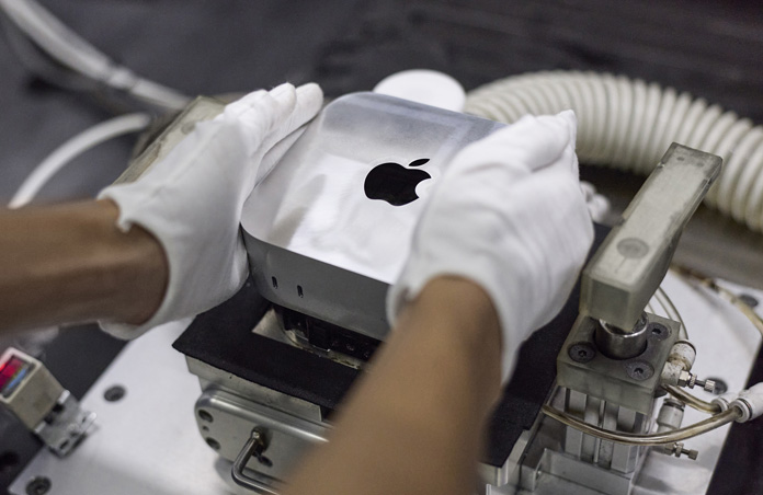 มือที่สวมถุงมือวาง Mac mini ลงในเครื่องจักรผลิตระหว่างการประกอบ